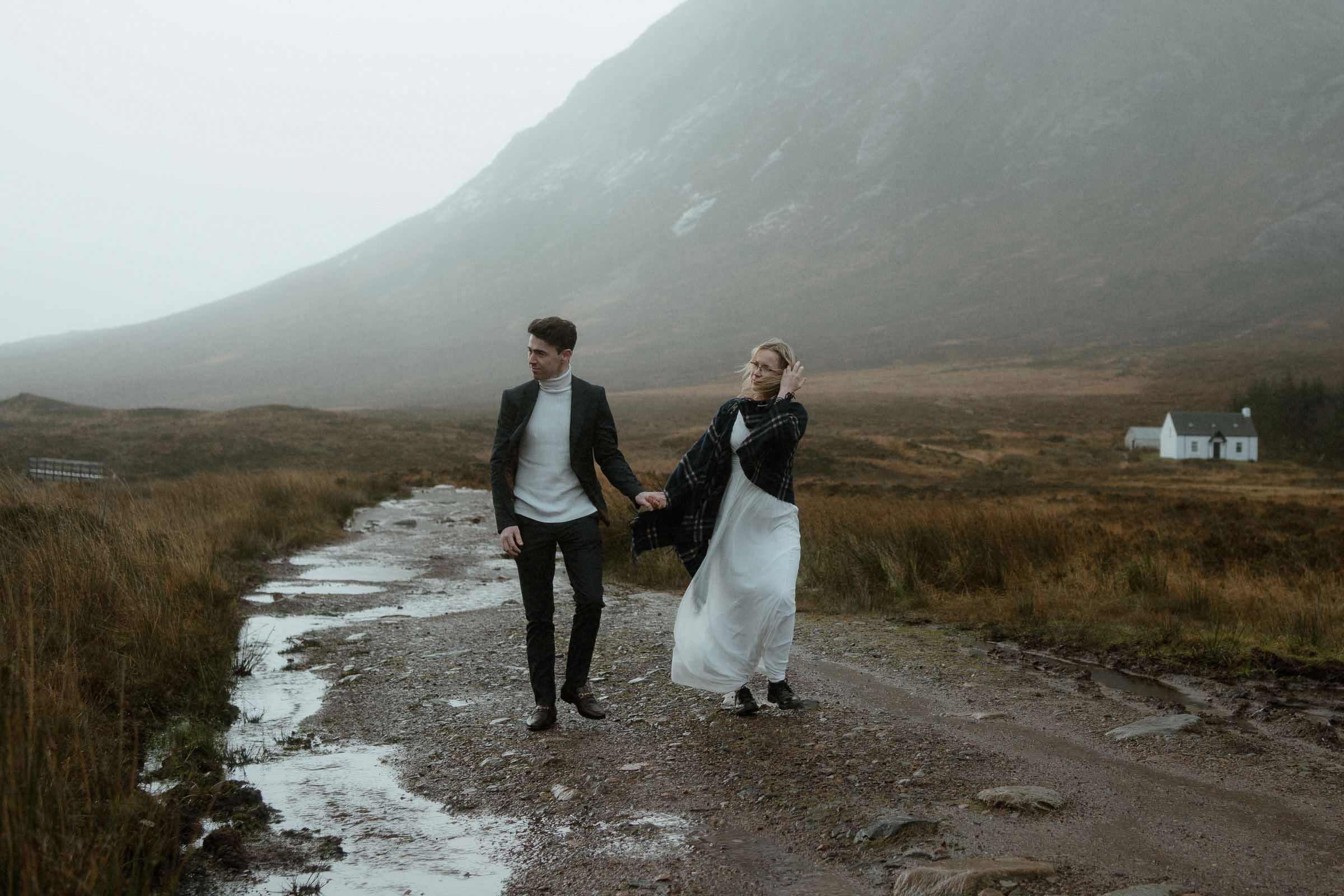 Couple hold hands and walk through Glencoe on a stormy day after their elopement in Scotland