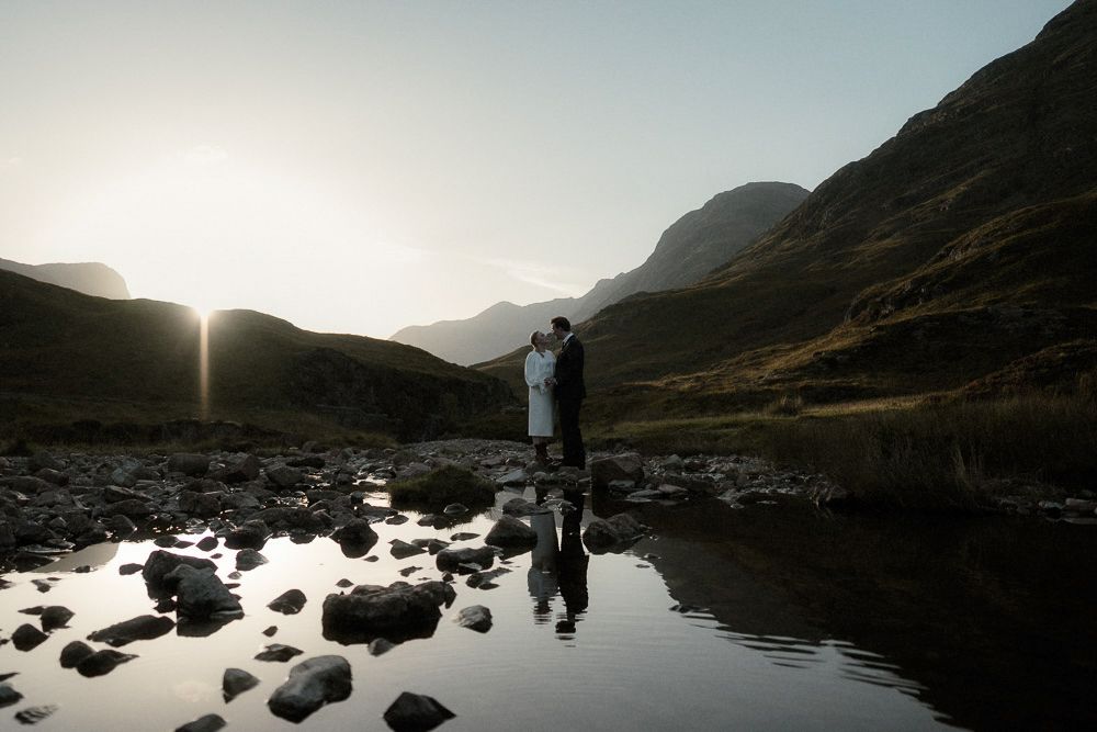 A newly-married couple stand on the banks of a loch during sunset after their elopement in Glencoe, Scotland