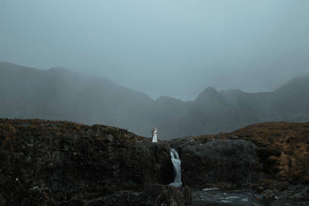 A couple hold hands by a waterfall with mountains in the background, after their elopement on the Isle of Skye, Scotland
