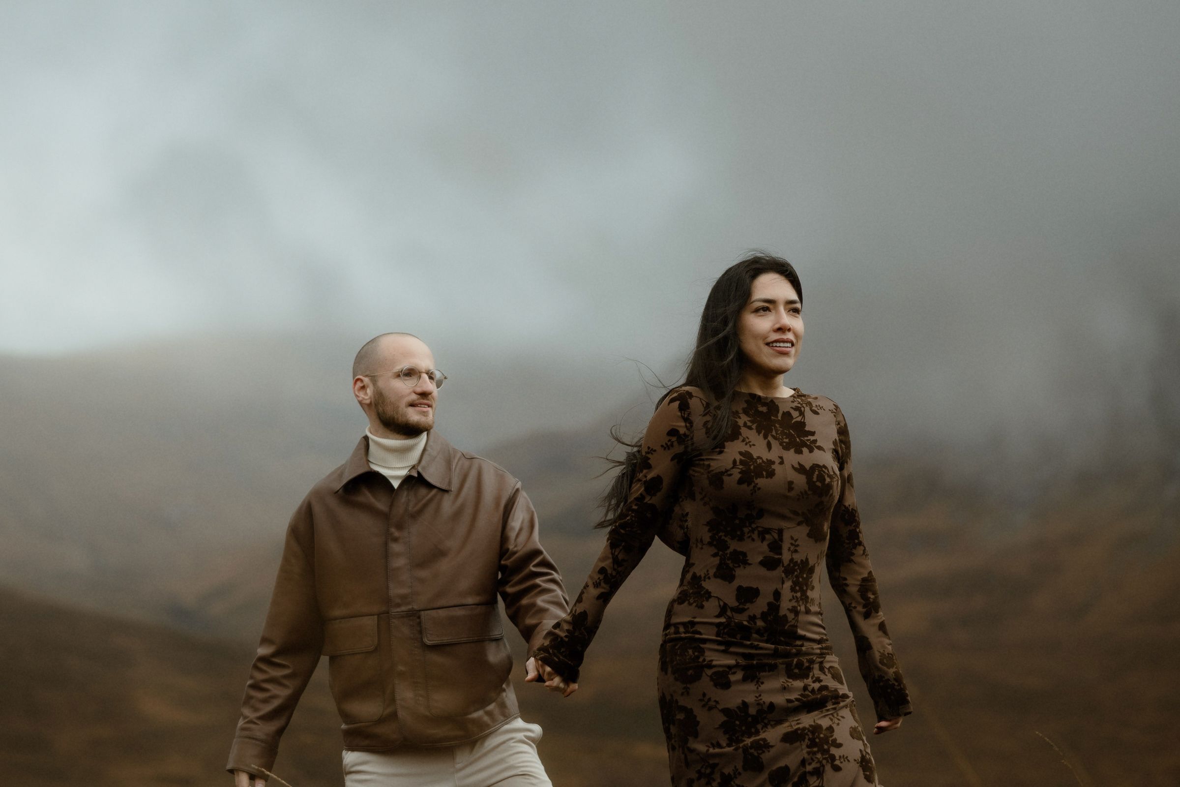A couple walk hand-in-hand in front of the misty mountains of Glencoe, Scotland
