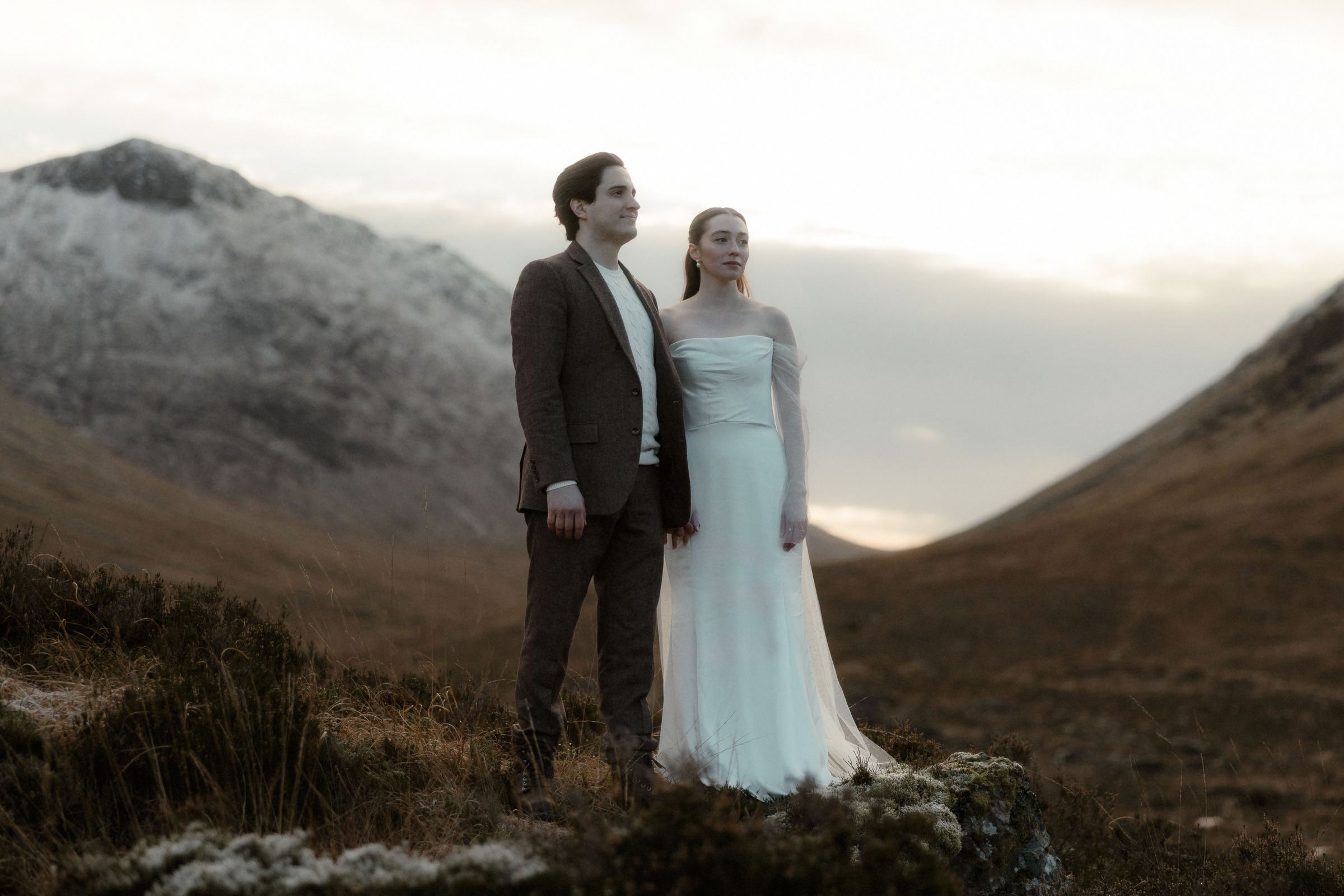 A newly-wed couple stand in front of the snowy mountains of Glencoe during their Scottish elopement photoshoot