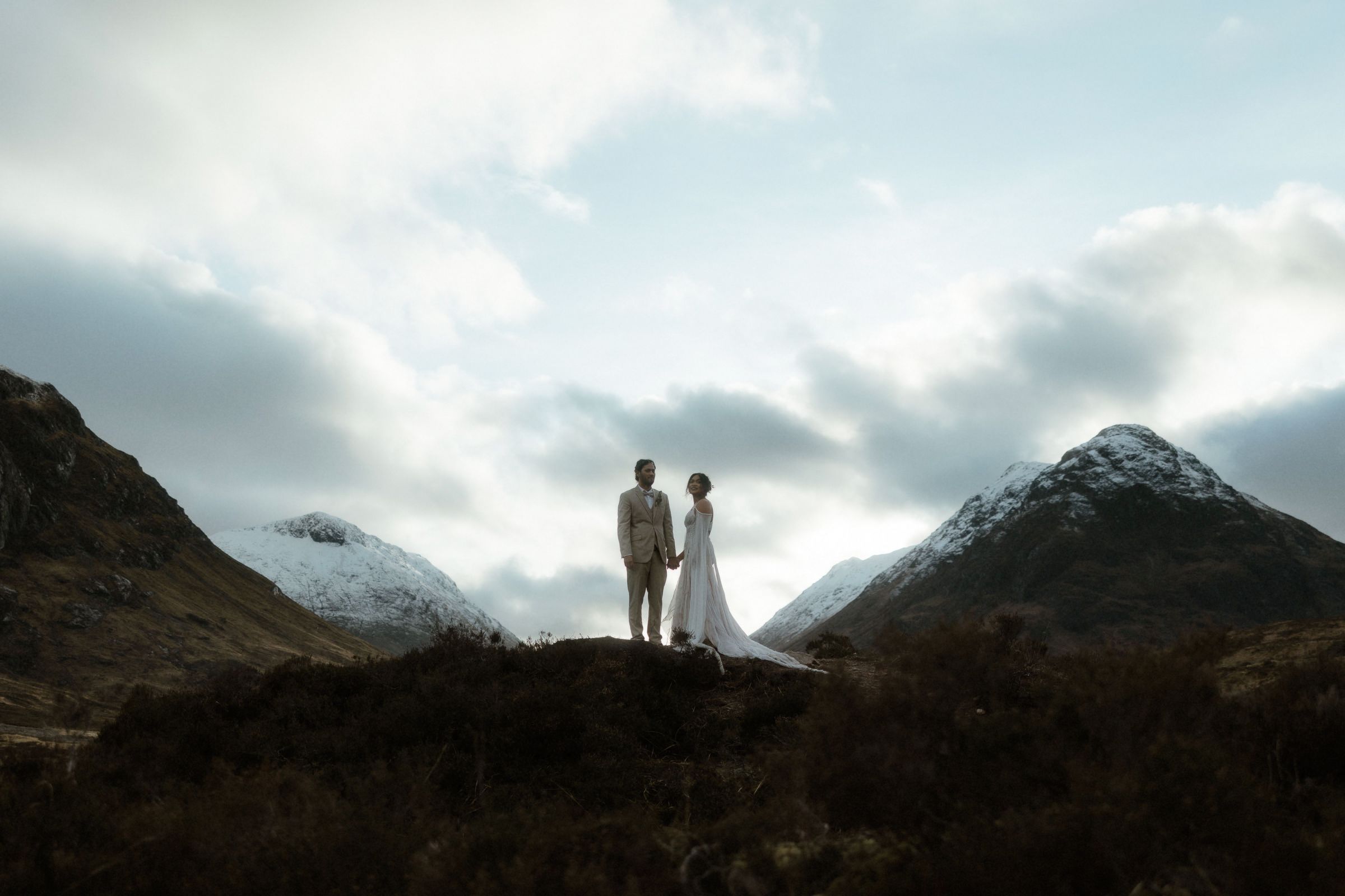 A bride and groom stand in front of snow-capped mountains during their winter elopement in Glencoe, Scotland