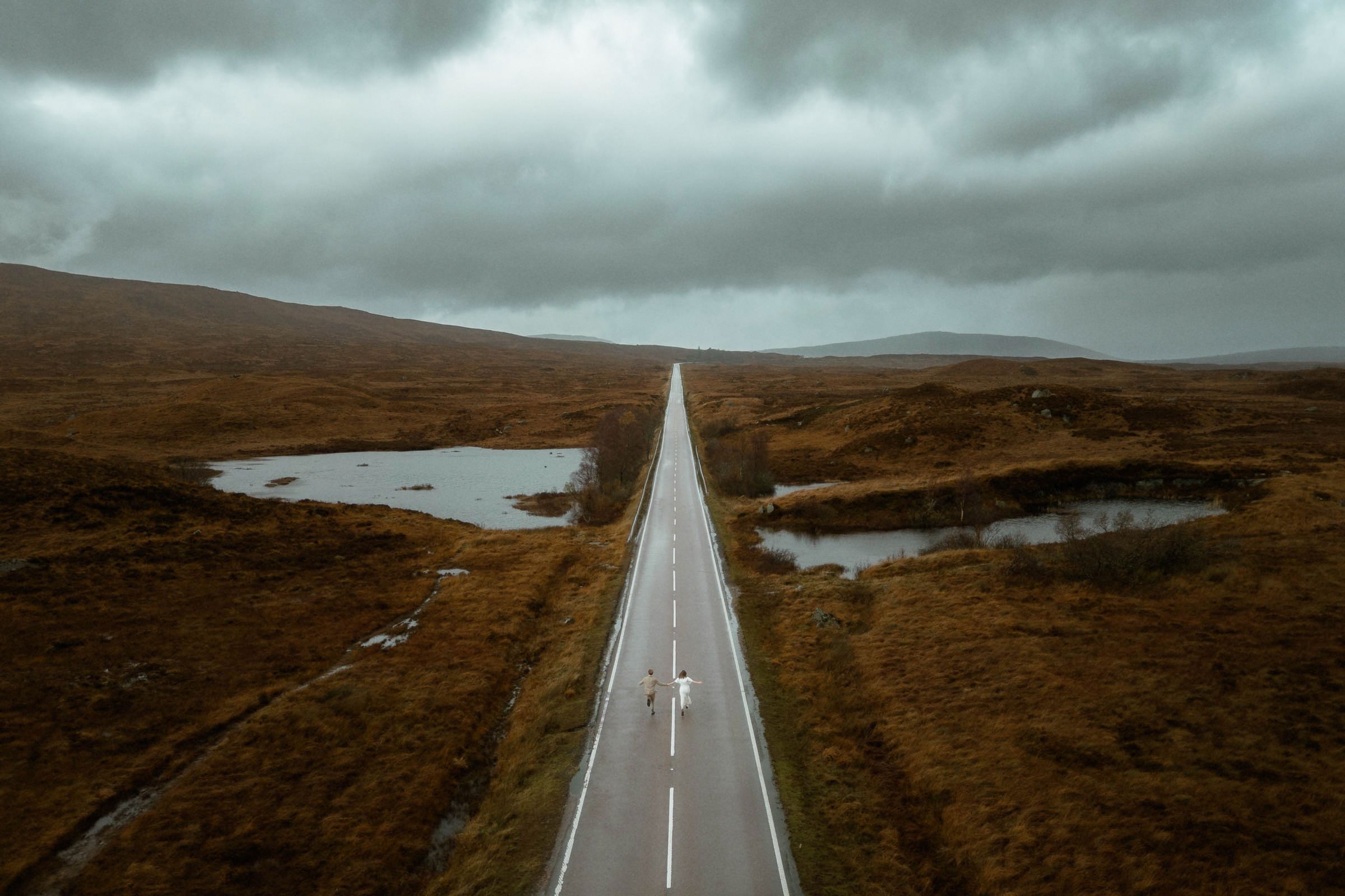 A bride and groom run up the middle of the road of Glencoe during their elopement in Scotland